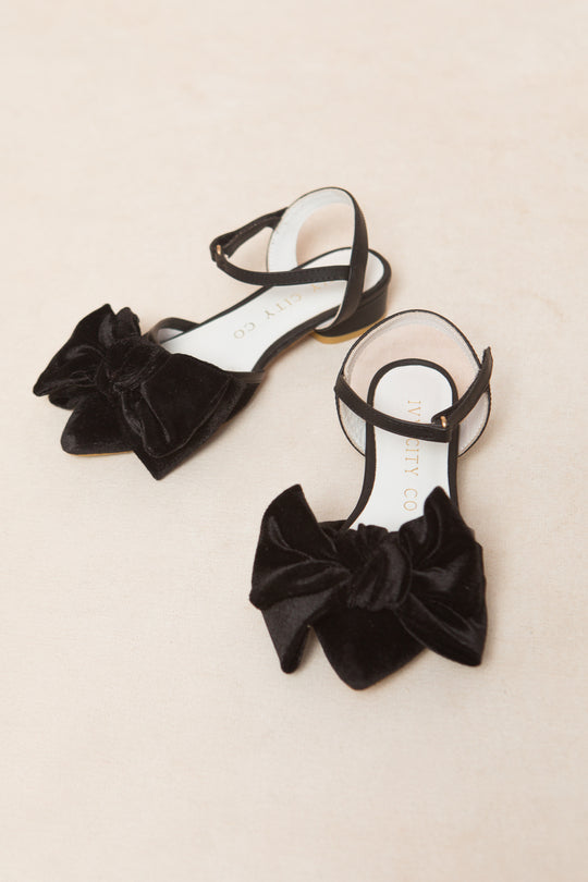 The Ivy City Co Mini Eloise Black Bow Heel features black velvet sandals with ankle straps and large bows on the toes, displayed on a light beige surface.