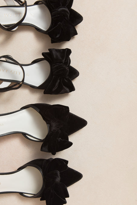Four pairs of Ivy City Co Mini Eloise Black Bow Heel flats with pointed toes and large velvet bows are neatly arranged on a light beige surface, displaying subtle gold branding on the black velvet insoles.