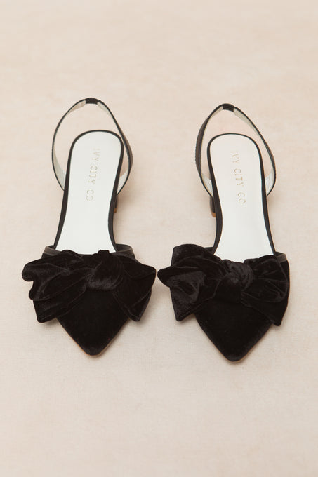 The Eloise Black Bow Heel by Ivy City Co features black pointed-toe slingback flats with large velvet bows, elegantly displayed on a light beige surface.