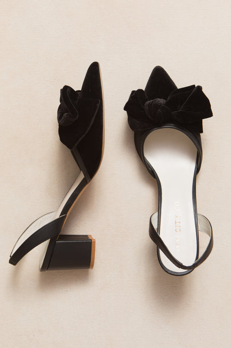 The Eloise Black Bow Heel by Ivy City Co features black velvet, pointed toes, block heels, and large decorative bows. Displayed on a light beige surface, these chic sling back shoes add elegance to any outfit.
