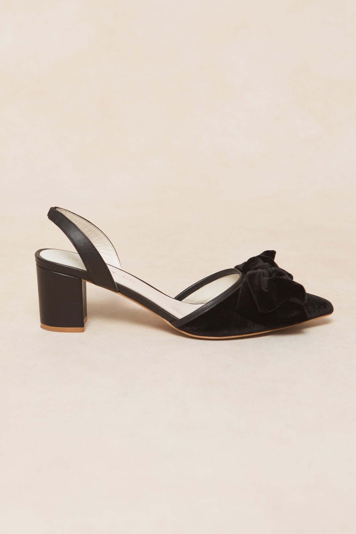 The Eloise Bow Heel in Black by Ivy City Co is a black velvet slingback with a closed pointed toe, large front bow detail, and mid-height block heel, shown against a light beige background.
