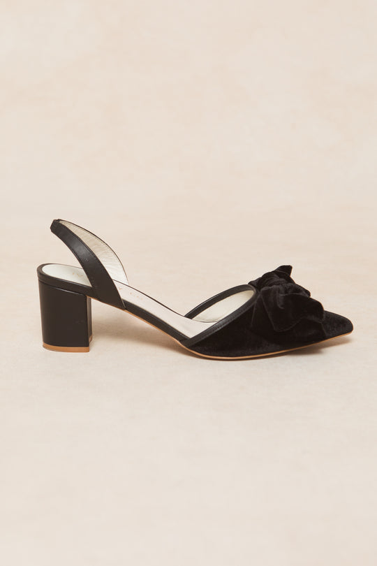 The Eloise Black Bow Heel by Ivy City Co is a black velvet slingback with a low heel, pointed toe, and elegant black bow detail on the front, shown against a neutral background.