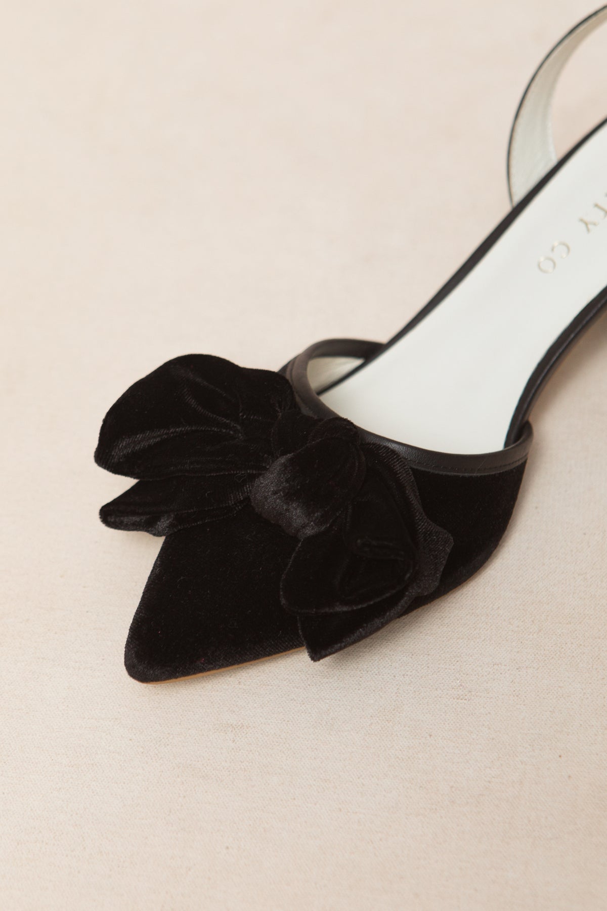 A close-up of the Ivy City Co Eloise Bow Heel in Black, a black velvet pointed-toe shoe with a large matching bow on the front, perfect for lovers of elegant black heels, displayed on a light beige surface.