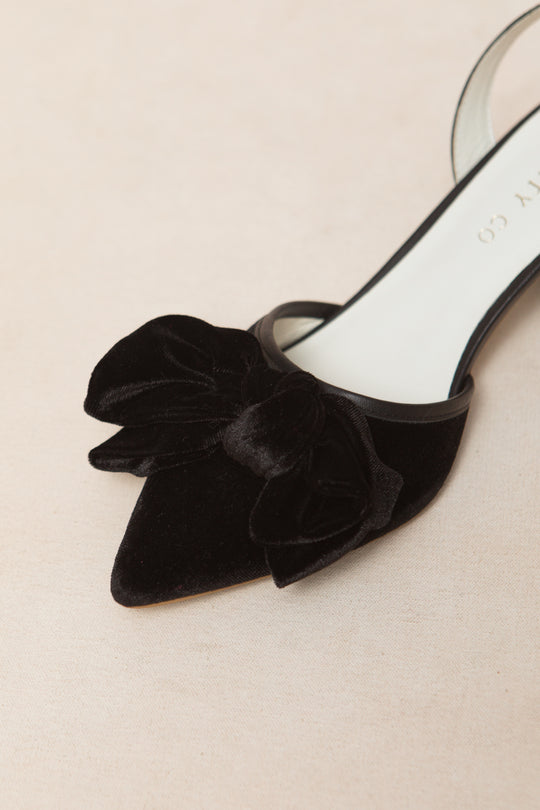 The Eloise Black Bow Heel by Ivy City Co is a black velvet pointed-toe slingback with a large front bow, shown against a light beige background.