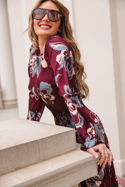 A woman wearing the Ivy City Co Elyse Dress in Burgundy Floral and large sunglasses smiles as she leans against a light-colored stone railing outdoors.