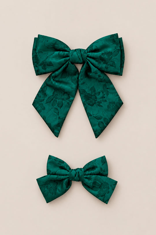 Two Hair Bows in Emerald Satin by Ivy City Co are displayed on a light beige background—one large and one small—arranged vertically with the larger bow above the smaller one. Their rich green floral pattern makes them perfect for use as an emerald hair bow or girls Christmas bow, and they are an ideal holiday photo accessory.
