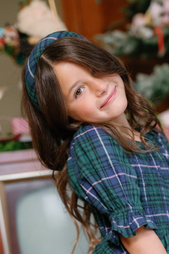 A young girl with long brown hair smiles and tilts her head, wearing Ivy City Co’s Hair Bows in Emerald Plaid as a festive accessory alongside a green plaid dress, set against a blurred backdrop of flowers and a vintage television.