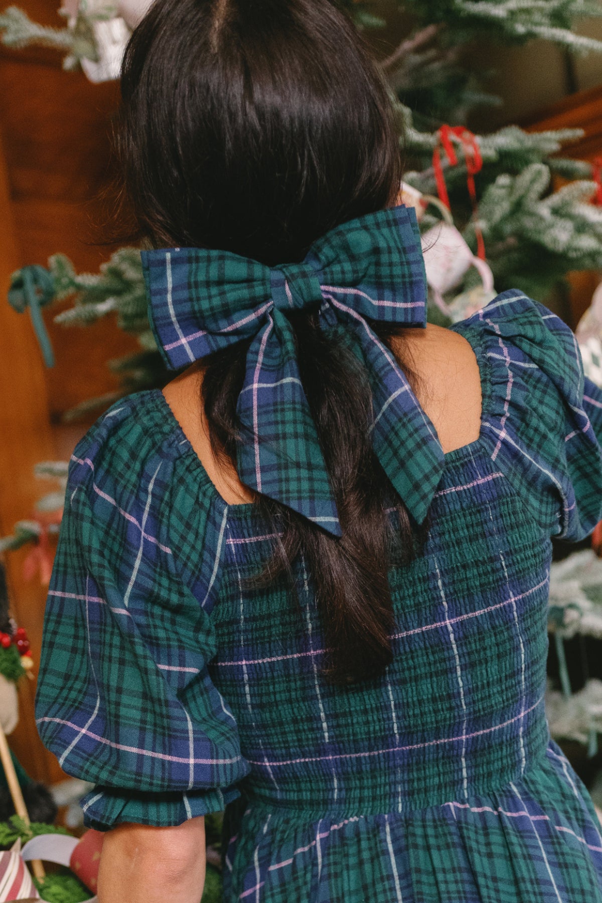 A person with long dark hair wears the Ivy City Co Hair Bows in Emerald Plaid and a matching plaid dress, standing in front of a decorated Christmas tree.