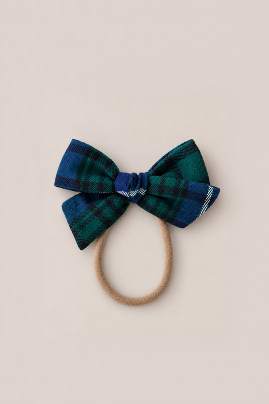 The Ivy City Co Hair Bows in Emerald Plaid feature a green and blue plaid bow attached to a tan elastic hair tie, shown on a simple light background—perfect as a festive holiday hair accessory.