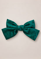 The Ivy City Co “Hair Bows in Emerald Satin” features a large emerald satin bow with a subtle floral pattern, perfect as a girl’s Christmas hair accessory or for holiday photos, beautifully displayed against a plain, light beige background.