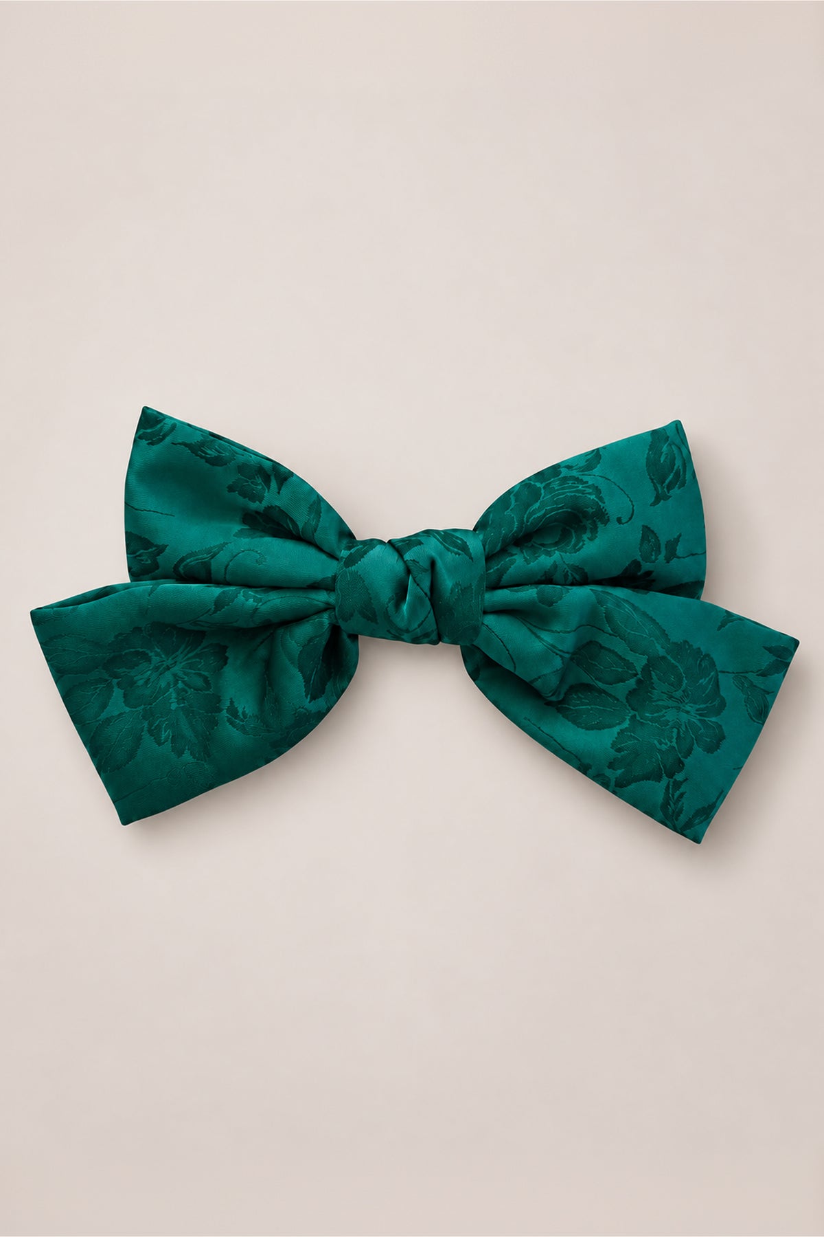 The Ivy City Co “Hair Bows in Emerald Satin” features a large emerald satin bow with a subtle floral pattern, perfect as a girl’s Christmas hair accessory or for holiday photos, beautifully displayed against a plain, light beige background.