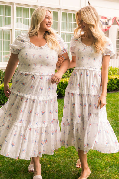 Two women smile at each other outdoors, both wearing the Ivy City Co Emily Dress in Heritage Soft Ditsy Floral—an ideal pick for a summer brunch. They stand on grass near a white house with large windows and leafy shrubs creating a charming backdrop.