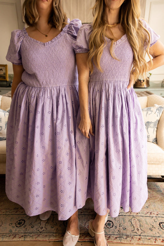 Two women stand side by side indoors, wearing the Emmi Dress in Lilac by Ivy City Co. These matching dresses feature short sleeves, puffed shoulders, and delicate eyelet cotton with intricate embroidered patterns. Their faces are not visible in the image, and the flattering silhouette is maternity friendly.