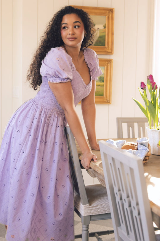 A woman with curly hair in the Emmi Dress in Lilac by Ivy City Co leans on a dining table adorned with flowers, bread, and napkins. The softly lit room features framed artwork on the wall behind her.