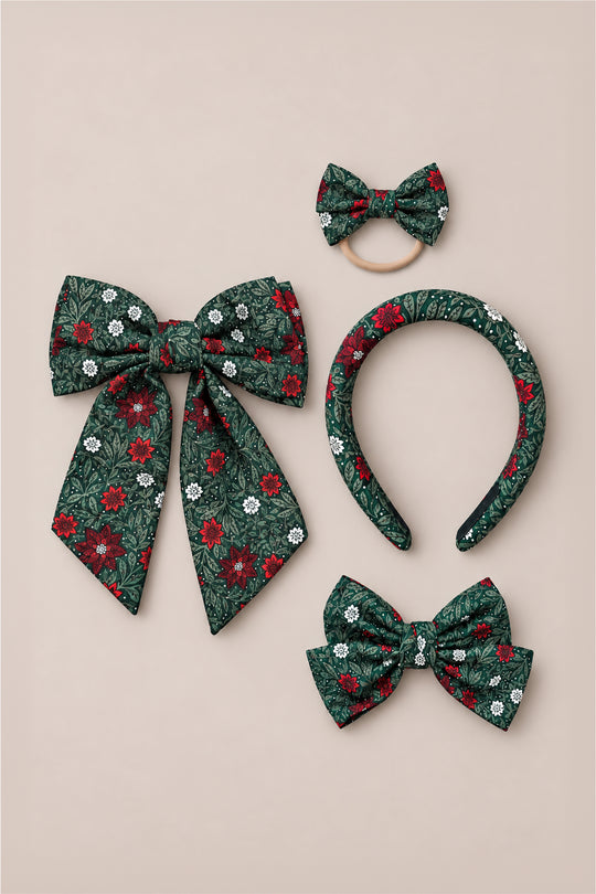 Four Ivy City Co “Hair Bows in Evergreen Merry Ditsy” hair accessories—featuring green, red, and white ditsy floral patterns—include a large bow, a small bow on an elastic band, a cotton headband, and a medium bow hair clip, all arranged on a beige background.