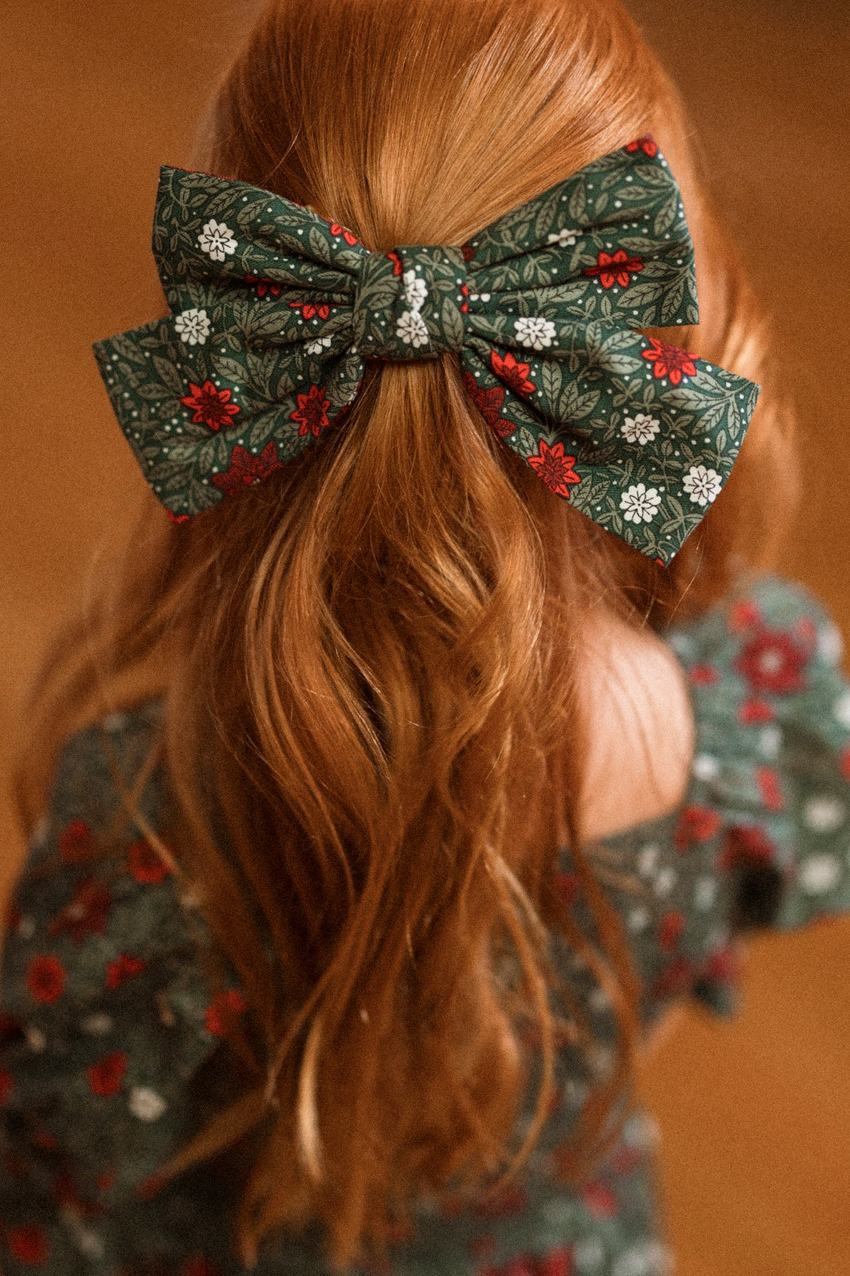 A person with long, wavy red hair is wearing the Ivy City Co “Hair Bows in Evergreen Merry Ditsy,” which coordinates with their green dress featuring a red and white floral pattern. The photo is taken from behind.