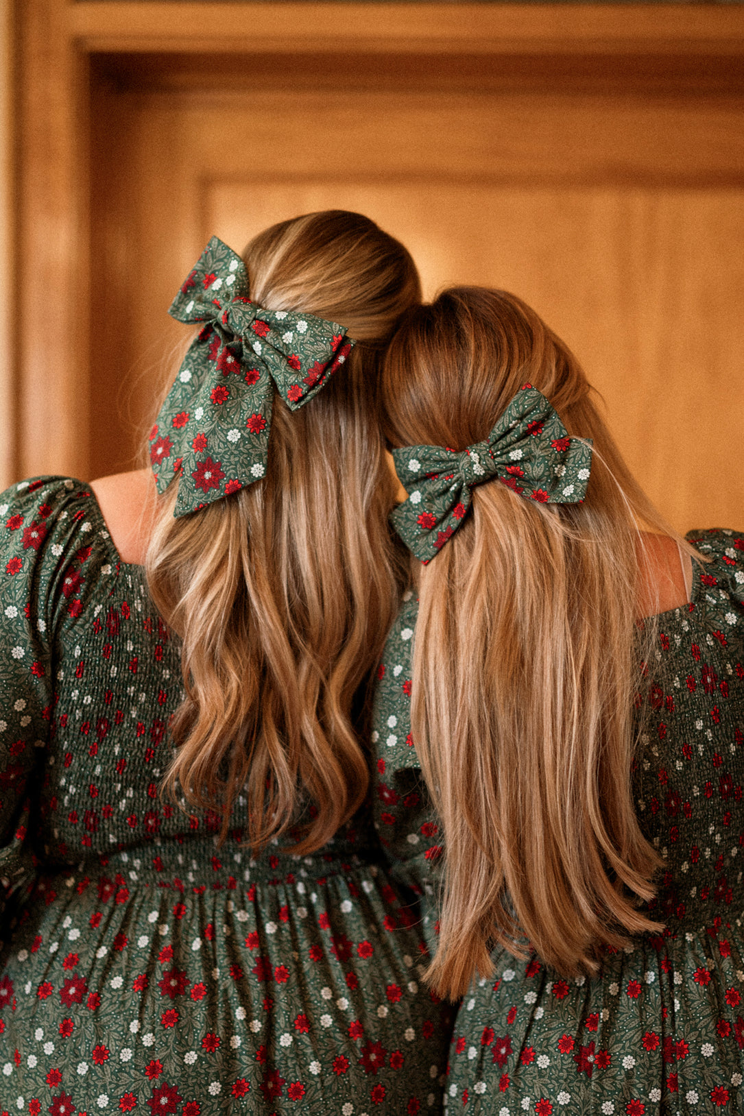 Two girls with long blonde hair, standing side by side with their backs to the camera in front of a wooden door, wear matching green ditsy floral dresses and Ivy City Co Hair Bows in Evergreen Merry Ditsy.