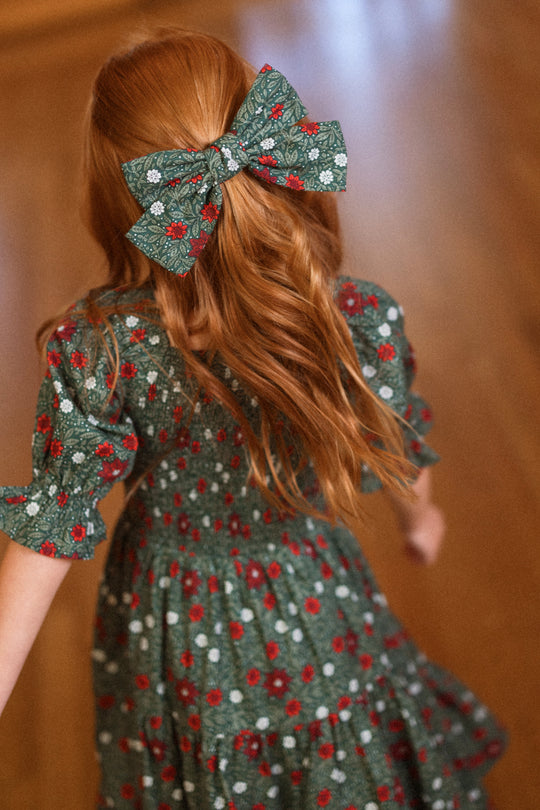 A young girl with long red hair wears an Ivy City Co Hair Bow in Evergreen Merry Ditsy, paired with a matching green ditsy floral dress, as she walks away on a wooden floor.