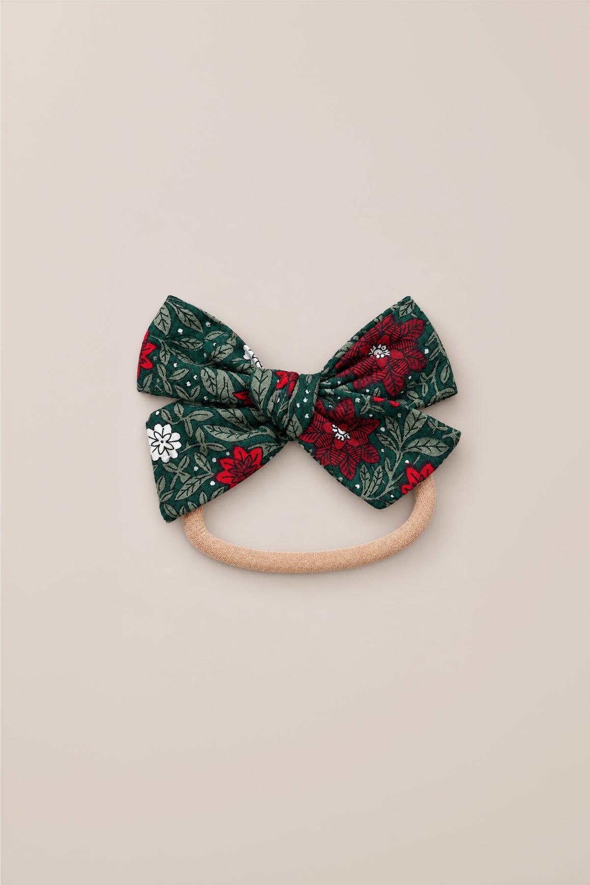 The “Hair Bows in Evergreen Merry Ditsy” by Ivy City Co features a green fabric bow adorned with ditsy floral patterns in red, white, and blue, attached to a light brown elastic band and displayed on a plain beige background.