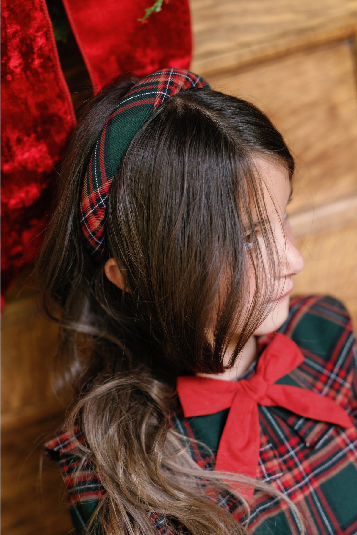 A young girl with long brown hair wears Ivy City Co’s Hair Bows in Evergreen Plaid, paired with a matching plaid cotton flannel dress, as she sits indoors near wooden paneling and red velvet fabric.