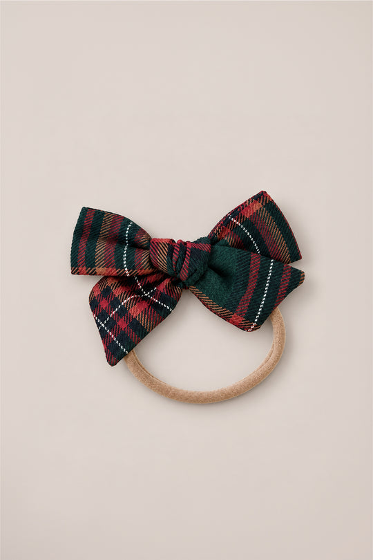 The Hair Bows in Evergreen Plaid by Ivy City Co feature a charming dark green, red, and gold cotton flannel bow attached to a beige elastic hair tie, shown on a simple light background—perfect as a festive holiday headband accessory.