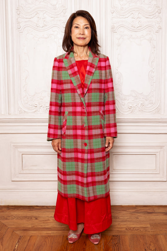 Woman wearing a red and green plaid coat over a red dress against a white wall.