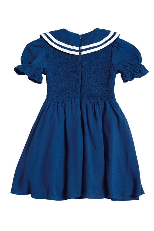 The Mini Alice Dress in Navy by Ivy City Co features a blue short-sleeve design with a white sailor-style collar, smocked bodice, ruffled sleeves, and a flared skirt. Shown from the back, it’s perfect for creating adorable Mom and Me matching looks.