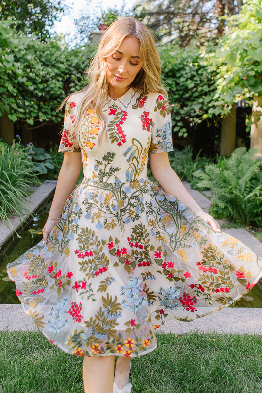 A woman with long blonde hair stands outdoors, holding the sides of her flowy Penelope Dress by Ivy City Co, which features a lace-trimmed neckline and colorful floral embroidery. Lush green plants and a small water feature are visible in the background.