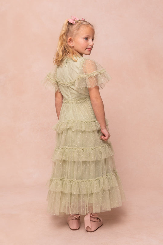 A young girl with blonde hair in a ponytail wears the Ivy City Co Mini Whimsical Maxi Dress in Sage—an elegant maxi-length gown with layered tulle skirt, ruffled accents, and sheer sleeves—paired with pink shoes as she stands slightly turned away against a soft pink background.