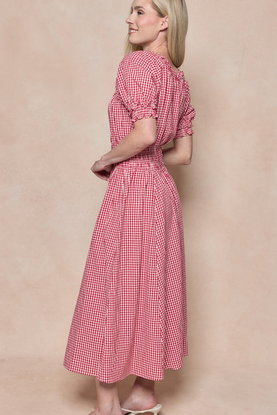 A woman with long blonde hair smiles as she stands sideways against a beige background, wearing the Ivy City Co Jessie Dress in Red Gingham with short puffed sleeves and white sandals.