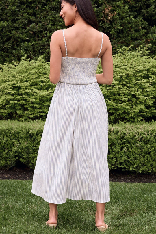 A woman stands on the grass, facing away, wearing the Ivy City Co Maxwell Dress in Blue & White Pinstripe—a light-colored, sleeveless, ankle-length nursing-friendly dress with thin straps. Green bushes are visible in the background.