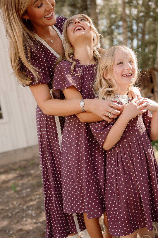 Three smiling and laughing—an adult woman and two young girls—enjoy time together outdoors in a sunlit, wooded area, all dressed in Ivy City Co’s Teen Blakely Dress in Aubergine Ditsy Dot with matching flutter sleeves.
