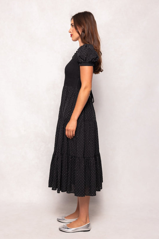 A woman with long brown hair stands sideways against a light neutral background, wearing the Ivy City Co Darby Dress in Black Diamonte—a maternity-friendly, short-sleeved midi dress adorned with small white polka dots—and pairs it with silver ballet flats.