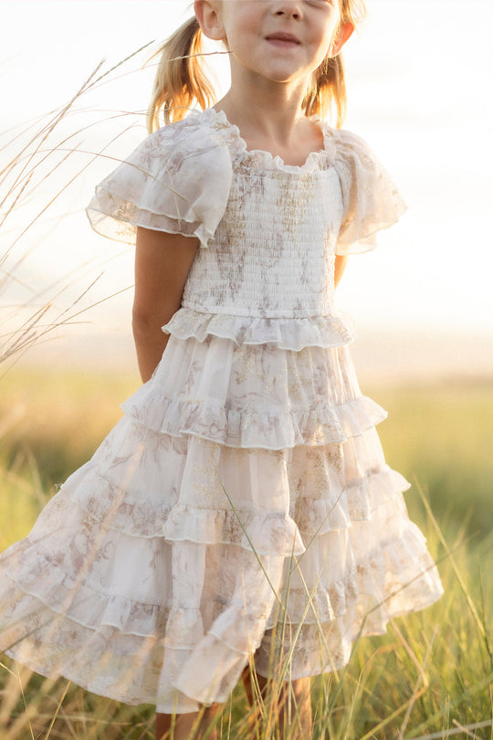 A young girl in a frilly, white, ruffled dress stands outdoors in tall grass, softly lit by sunlight. Her face is partially out of frame, and she has her hair in pigtails. The delicate details of her Ivy City Co Mini Winnie Dress in Bright White Wispy Wildflower lend a dreamy charm to the scene.