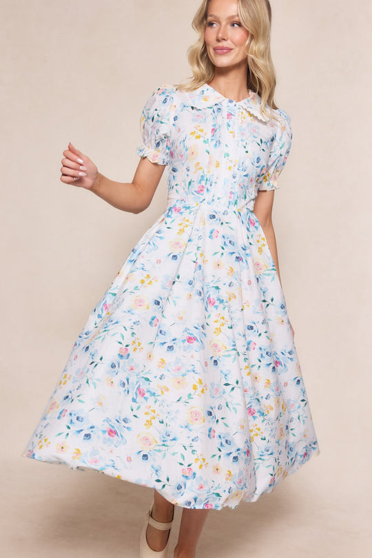 A woman models the Ivy City Co Sophia Dress in Blue Bouquet, a floral, midi-length piece with puff sleeves and a fitted waist, smiling softly against a neutral background.
