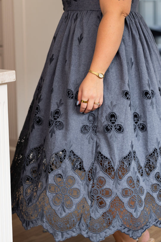 A person wearing the Ivy City Co Penny Tank Midi Dress in Embroidered Chambray, featuring gray cotton chambray fabric with floral eyelet lace detailing at the hem, along with a gold bracelet and two rings. Only the lower half of the dress and part of their arm are visible.