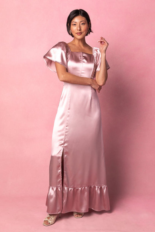 A woman stands against a pink background wearing the Ivy City Co Maddie Dress in Blush Satin, featuring flutter sleeves, a ruffle hem, and a thigh-high slit. She completes the elegant look with open-toe heels and earrings—perfect as a bridesmaid dress or for any special occasion.