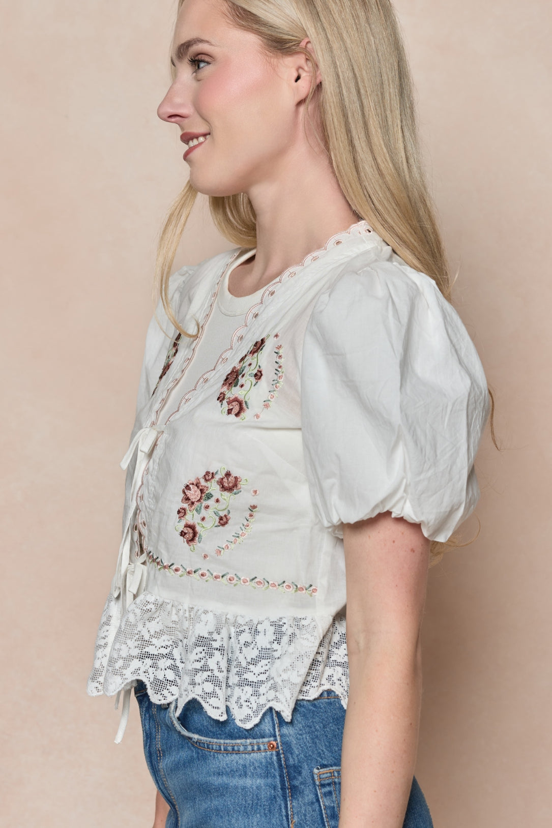 A woman with long blonde hair stands against a beige background wearing the Ivy City Co Fable Top in White—a peplum style with puff sleeves, scalloped lace hem, and floral embroidery—paired with blue jeans.
