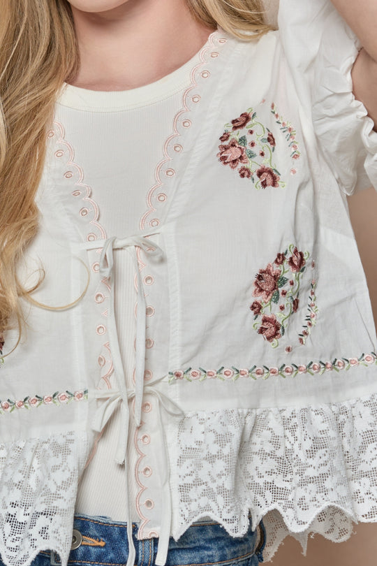 A close-up of a woman in the Ivy City Co Fable Top in White, featuring floral embroidery, scalloped lace hem, eyelets, and front tie details over a cream shirt, with her long blonde hair partially visible.