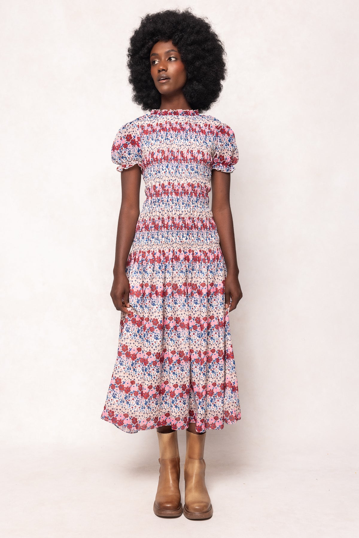 A woman with a natural afro wears the Ivy City Co Hayden Dress in Lush Garden—a romantic, smocked, short-sleeve, midi dress in red, blue, and white—with brown ankle boots, standing against a light background.