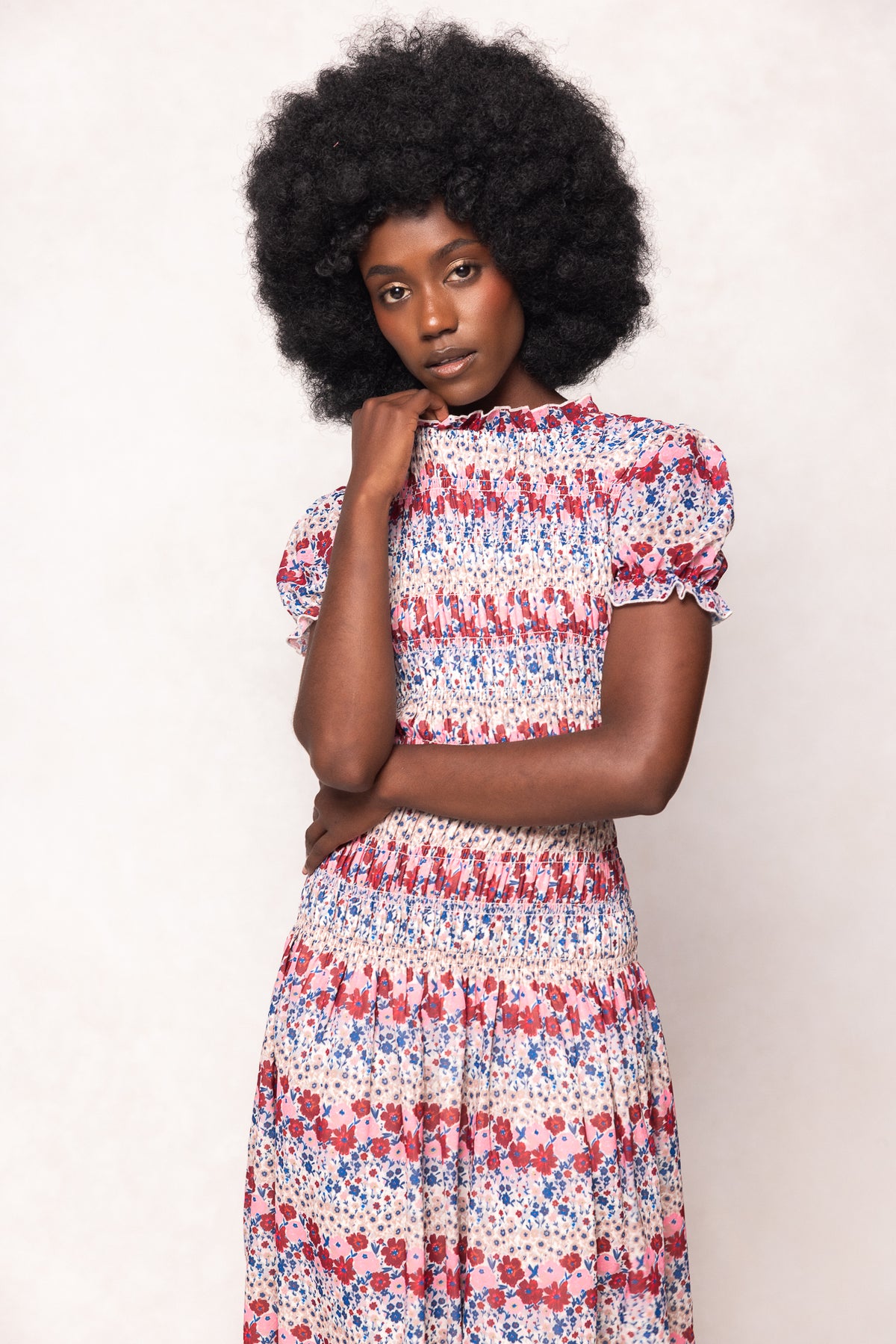 A woman with a natural afro poses against a light background, resting her chin on her hand and looking at the camera while wearing the Ivy City Co Hayden Dress in Lush Garden, featuring romantic red, blue, and white floral patterns.