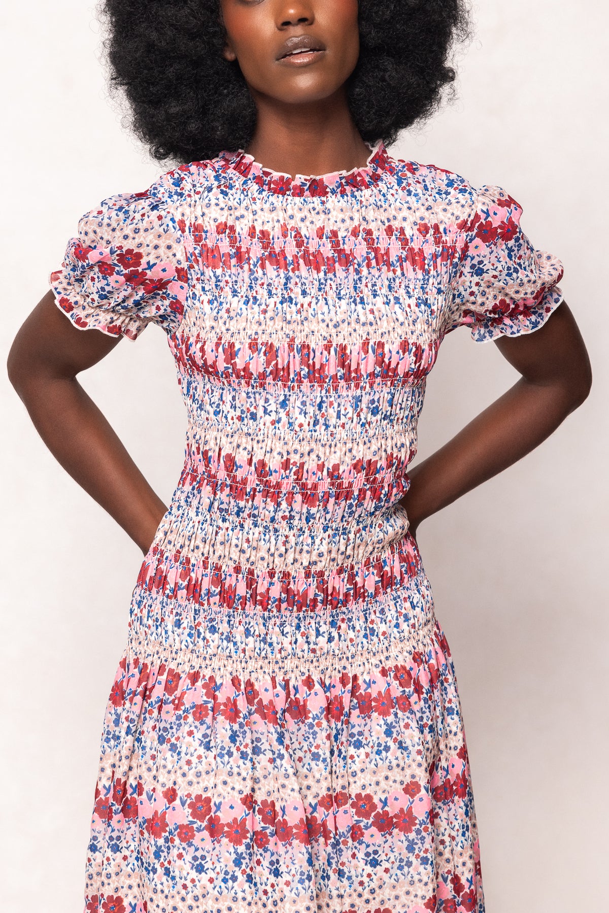 A woman wears the Ivy City Co Hayden Dress in Lush Garden, a romantic floral midi with short sleeves, ruffled edges, and a smocked bodice in red, blue, and pink. She stands against a light background with her hands behind her back.
