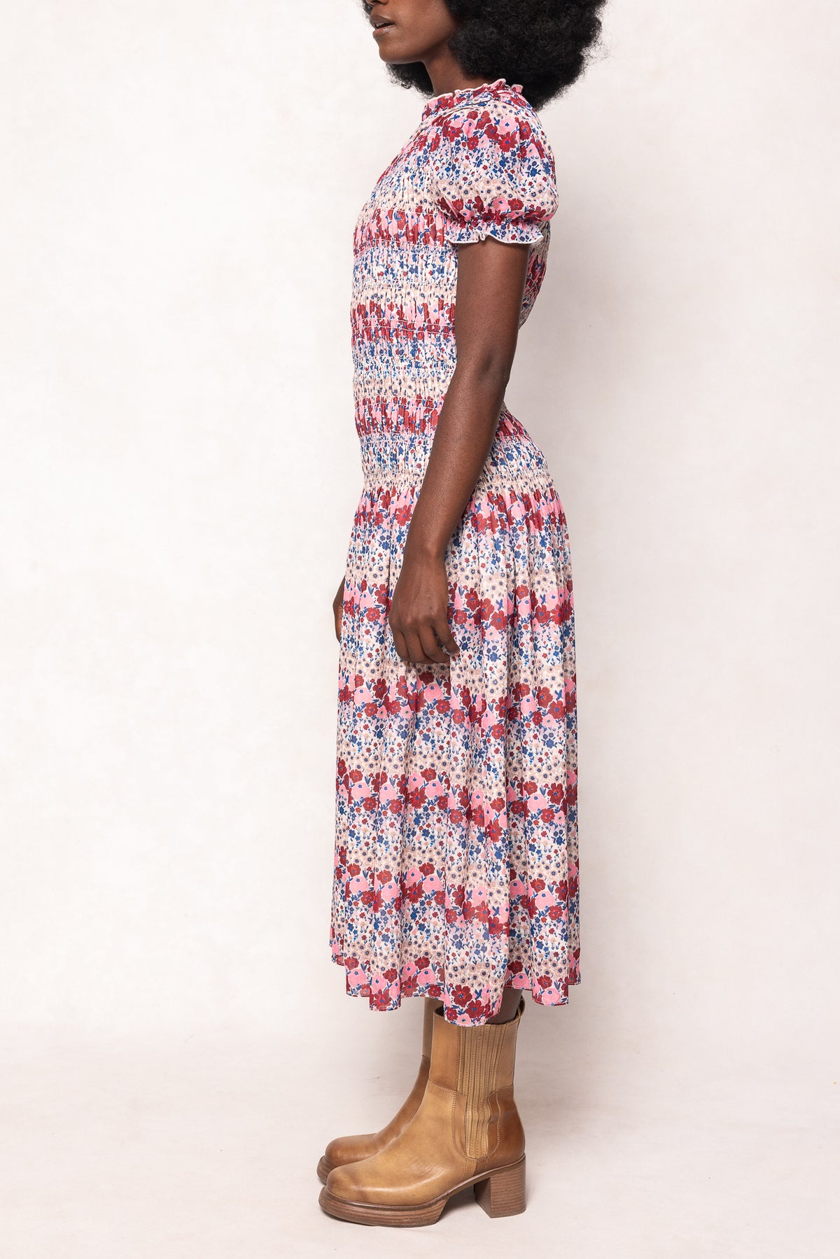 A woman stands in profile against a plain background wearing the Ivy City Co Hayden Dress in Lush Garden—a romantic, midi-length floral dress with shirred detailing—paired with light brown boots for an effortlessly chic look.