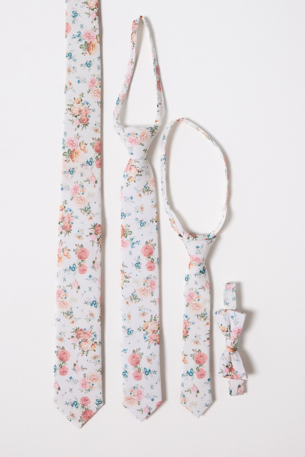 The Ties in Countryside Peony Blossom set by Ivy City Co features four matching accessories—adult, child, and baby neckties plus a family bow tie—in a pink, blue, and green floral pattern on white. All are displayed on a white background.