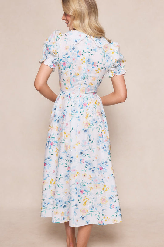 A woman stands with her back to the camera, wearing the Sophia Dress in Blue Bouquet by Ivy City Co—a light blue midi dress with puffed short sleeves, a fitted waist, and a pastel floral pattern.