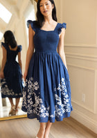 A woman stands indoors wearing the Ivy City Co Finch Dress in Blue, a sleeveless, knee-length cotton dress with white embroidery. Her reflection appears in a large mirror. She pairs the dress with white shoes and has long dark hair.