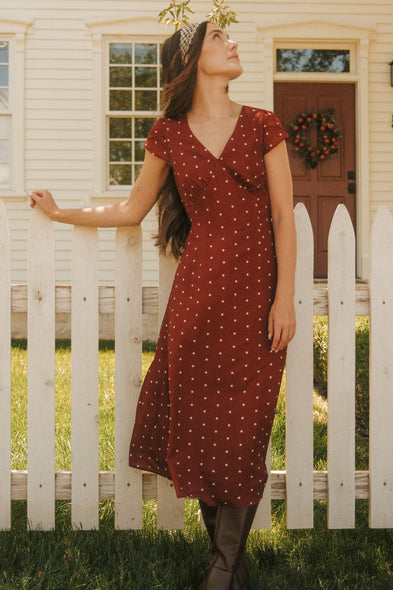 A woman wearing the Ivy City Co Fiona Dress in Cranberry Dainty Dot, featuring petal sleeves, pairs it with brown boots as she leans on a white picket fence in front of a cream-colored house with a wreath on the door—making this outfit the perfect addition to your fall wardrobe.