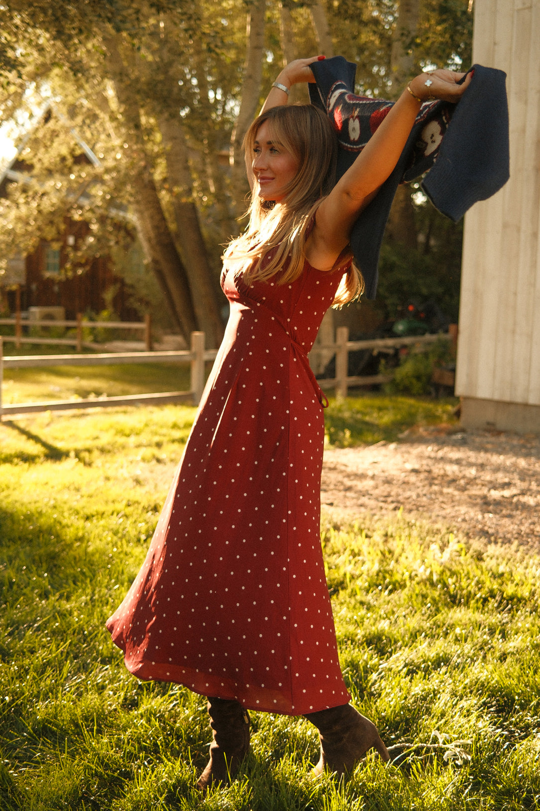A woman stands on the grass in Ivy City Co’s Fiona Dress in Cranberry Dainty Dot, featuring delicate petal sleeves. She lifts a dark jacket above her head as sunlight streams through the trees behind her, casting a golden glow—an inspiring scene for your fall wardrobe.