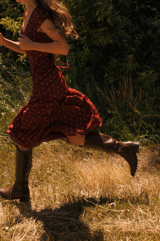 A person in the Fiona Dress in Cranberry Dainty Dot by Ivy City Co, featuring petal sleeves and paired with brown boots, is running through tall, dry grass with greenery in the background. Only the lower half of the body is visible—an effortless nod to a fall wardrobe.