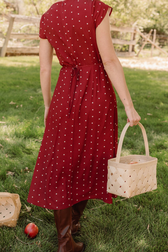 A person seen from behind, wearing the Ivy City Co Fiona Dress in Cranberry Dainty Dot with petal sleeves and brown boots, walks through a grassy area while carrying a light-colored woven basket. A single red apple lies on the grass near another basket—perfect inspiration for your fall wardrobe.